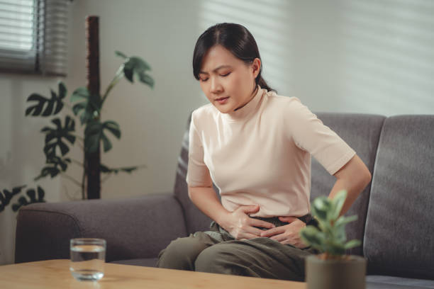 azjatka czująca ból brzucha siedząca na kanapie w salonie w domu. - peptic ulcer zdjęcia i obrazy z banku zdjęć