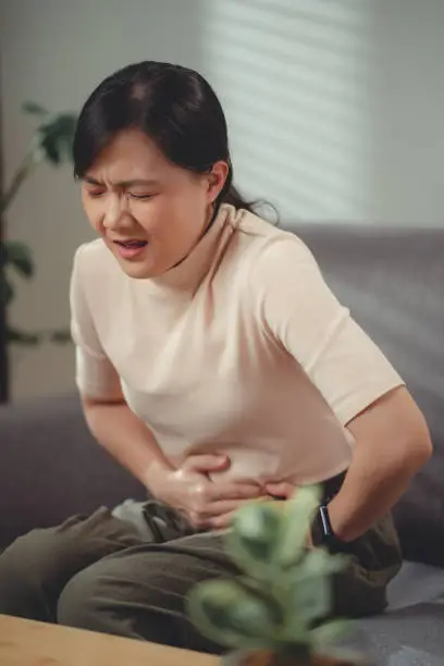 Photo of Asian woman feeling stomachache sitting on sofa in living room at home.