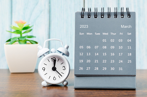 March 2023 Monthly desk calendar for 2023 year with alarm clock on blue wooden background.