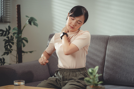 Asian woman suffering from neck pain massaging on occipital by hands sitting on sofa in living room at home.