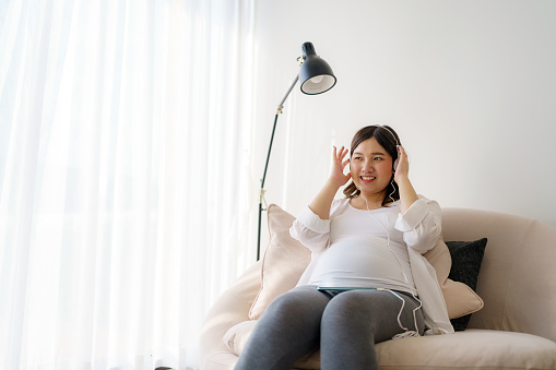 Portrait of pregnant Happy Asian woman sitting on sofa at home and using smart phone for listening music in headphones.