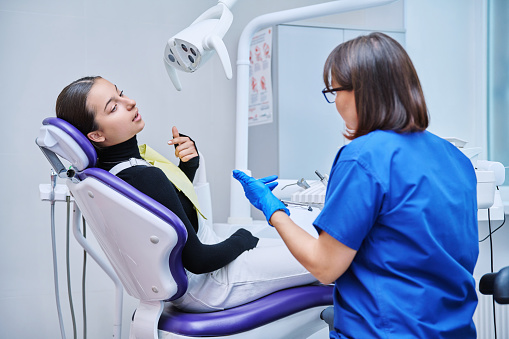 Dentist and teenage girl patient in dental clinic, doctor nurse writing on clipboard consulting, teen female talking about toothache disturbing dental problem. Dentistry, treatment, dental health care