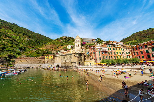 Vernazza, Italy - July 22th, 2019: Port and sandy beach of the ancient Vernazza village crowded with tourists on a hot and sunny summer day. Cinque Terre, National park in Liguria, La Spezia, Italy, Europe. Some tourists and locals sunbathe on the beach or swim in the village harbor. This natural park (UNESCO world heritage site) includes the villages of Riomaggiore, Manarola, Corniglia, Vernazza and Monterosso al Mare.