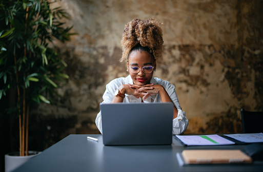 Smiling African-American woman reading business report on her laptop while sitting at restaurant desk.