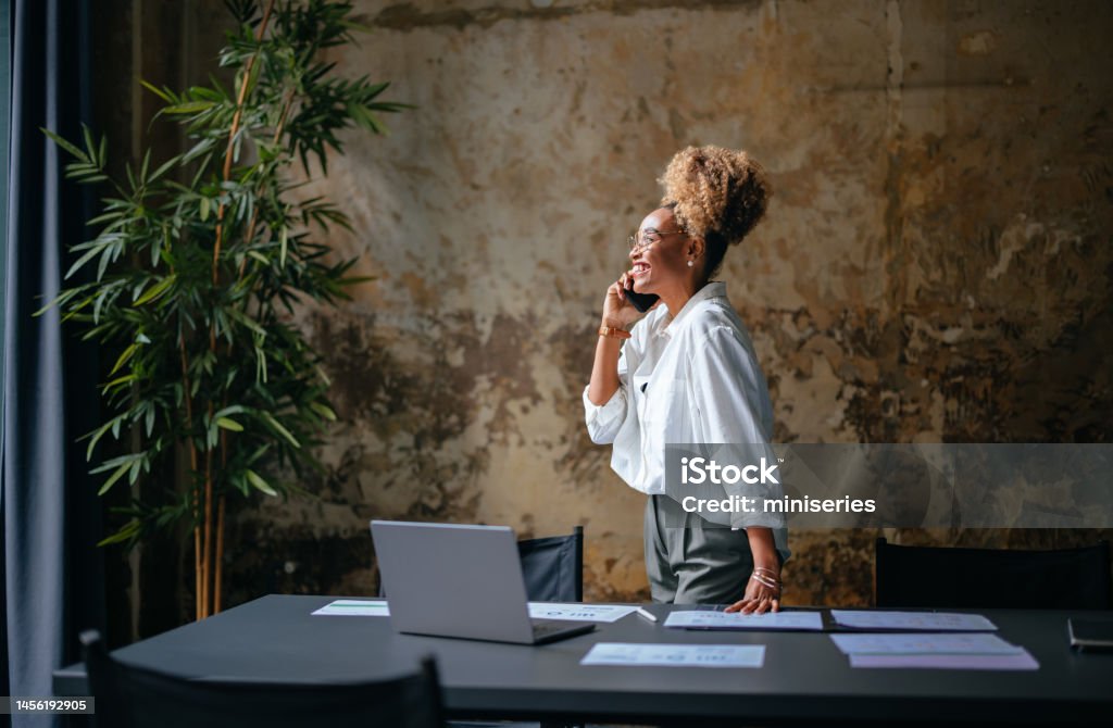 Smiling Businesswoman Talking On A Mobile Phone In The Cafe Happy African-American woman speaking on the phone while standing at restaurant desk with a laptop computer and documents. Entrepreneur Stock Photo