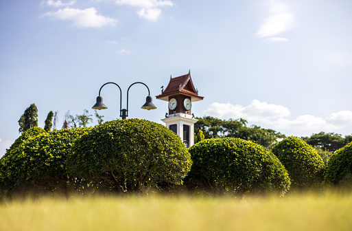 A low angle view looking through the grass blurs the foreground to round bushes and decorative lanterns and clock towers in a park with sky clouds as an afternoon backdrop.