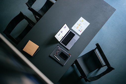 High angle view of desk and chairs with laptop computer, documents, mobile phone, notebook, pen and eyeglasses.