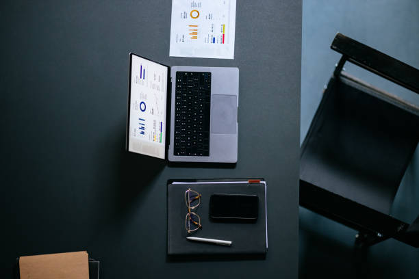 High angle view of desk and chair with laptop computer, documents, mobile phone, notebook, pen and eyeglasses.