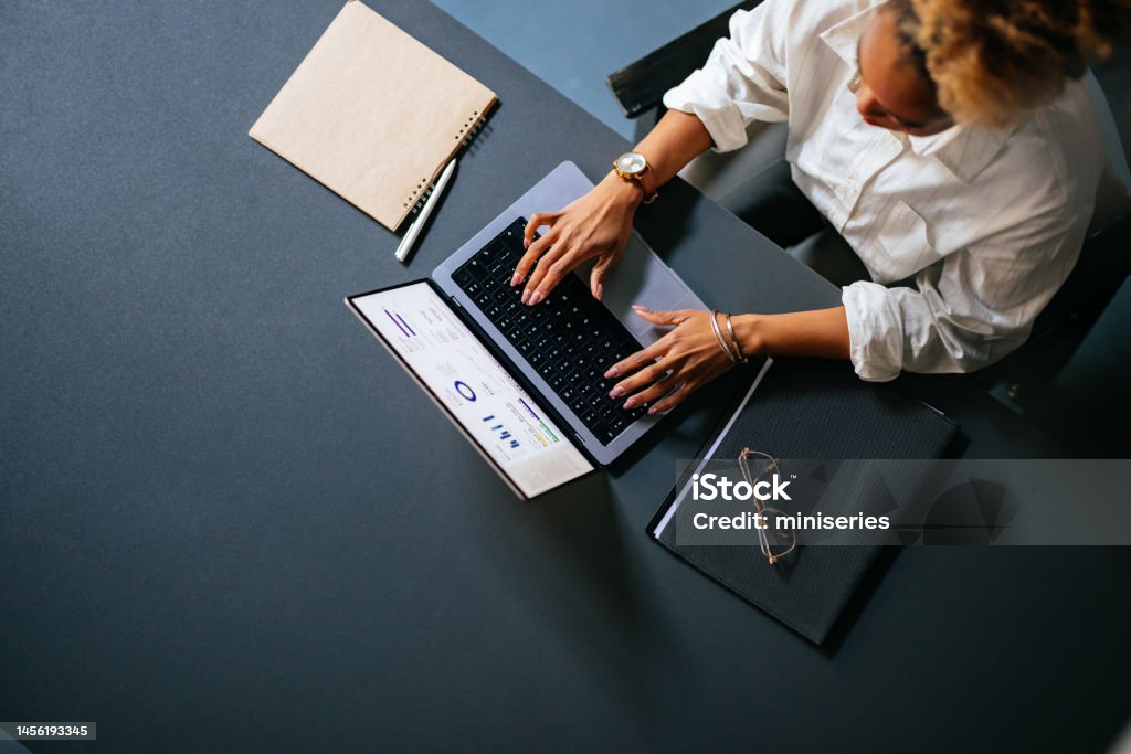 High Angle View Of Unrecognizable Woman Typing Business Report On A Laptop Keyboard In The Cafe From above photo of an anonymous African-American woman analyzing business graph on a laptop computer while sitting at restaurant desk with notebook, pen and eyeglasses. People Stock Photo