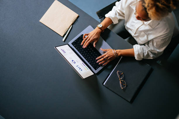 High Angle View Of Unrecognizable Woman Typing Business Report On A Laptop Keyboard In The Cafe From above photo of an anonymous African-American woman analyzing business graph on a laptop computer while sitting at restaurant desk with notebook, pen and eyeglasses. using computer stock pictures, royalty-free photos & images