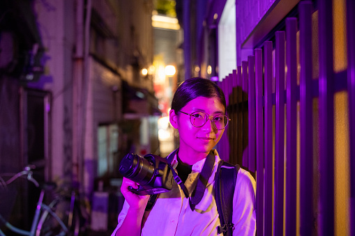 Portrait of female tourist standing on narrow footpath in purple light at night, holding digital camera