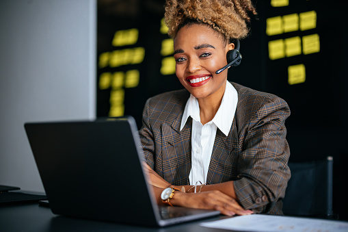 Happy African-American woman with wireless headset working in customer service with her laptop computer while sitting at office desk and looking at camera.