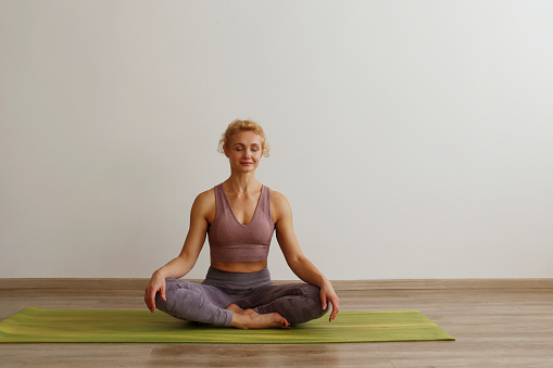 Adult sporty woman practicing yoga indoors. Yogini sitting on green mat and meditating at home. Close up, copy space, background.