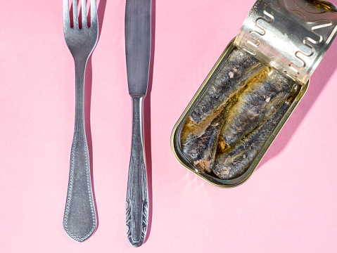 Tinned sardines on olive oil, knife and fork isolated on pink background