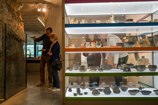 Two heterosexual couples at the museum, full length shot of one admiring the ruins of a Roman wall, the other couple observing at the kitchenware made of clay. Strong light contrasts.