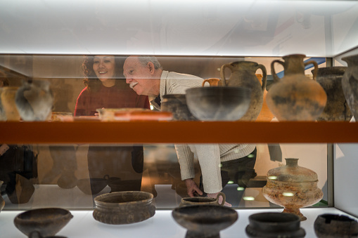 Shot through the glass shelves, focus on a pretty  woman and her senior male friend at the exhibition of ancient clay works. Both looking away and having fun. Ancient bowls in the foreground, waist up image.