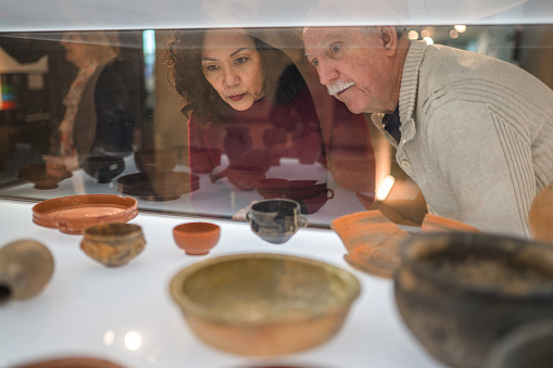 Shot through the glass, a close up of a beautiful mature woman and her senior male friend at the exhibition of ancient terracotta pots. Both looking away. Lots of different pottery in the foreground.