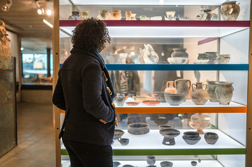 Well lit glass display showing various earthenware, a black-haired man standing in front of the shelves, looking at the objects. Only back view of the man, contrasting light.