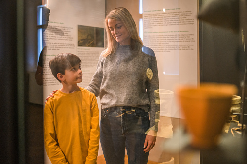 A Caucasian mother with school aged son  visiting a history museum and admiring the objects in display cases. 3/4 length image, shot through the glass, both looking away.