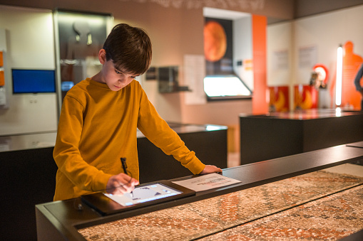 A school-aged boy in sweat shirt using an interactive museum label at the history museum, a Roman mosaic in front of the boy. Waist up image.