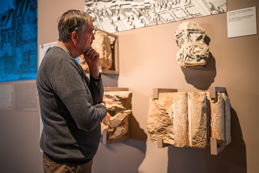 A senior man in casual clothes standing at the museum, his hand supporting the other elbow while he is carefully examining the stone objects found in an ancient Roman town.