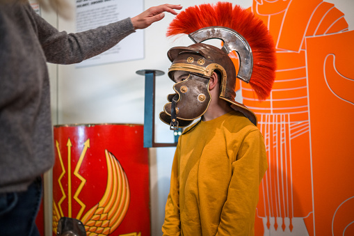 School-aged boy trying on a soldier's helmet at an exhibition of Ancient Roman Empire. Wall paintings of Roman legionaries in the background. No face visible.