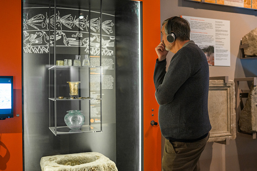 Senior Caucasian male museum visitor listening to an audio guide, using headphones while looking at objects from Roman era. 3/4 length image, back view.