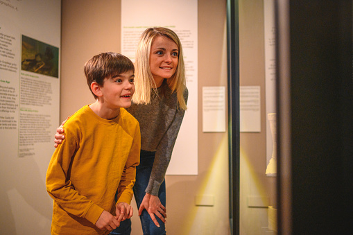 Close up of a pretty woman and her school-aged little brother amazed by the artifact exhibited in a glass case. Looking away, 3/4 length image.