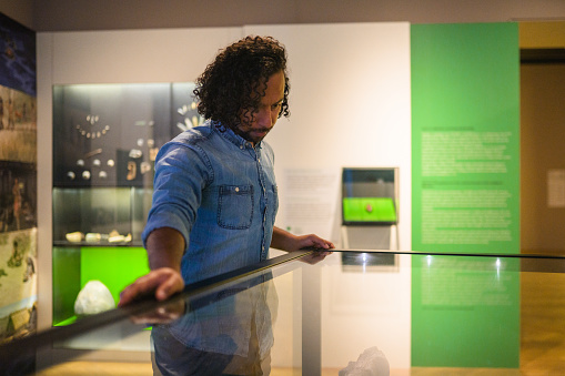 Handsome mid adult male museum guest looking at the objects exhibited under glass, his reflection visible in the glass. Waist up image, various objects and information boards in the blurred background.