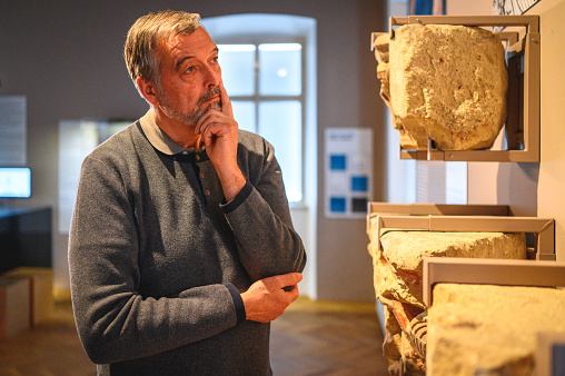 A male museum visitor of senior age, reading the data of an artifact at a history museum. Waist up image, spotlight on the stone objects. Looking away, his palm supporting the chin.