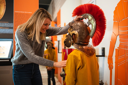 Young female Caucasian museum employee fixing a helmet of a Roman legionary on the head of a little boy. Back view of the boy. Various descriptions and a screen display on the walls in blurred background.