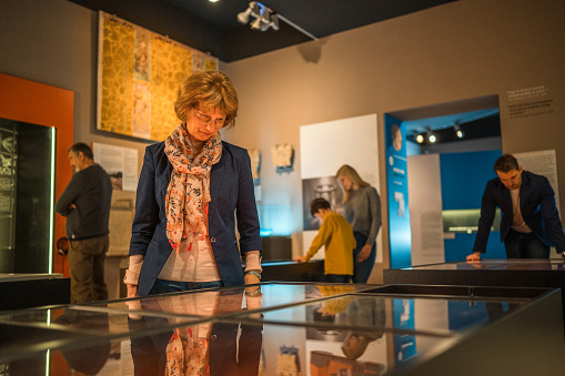 An elegant Caucasian woman looking at the museum display cases. Lots of people in the background, waist up shot over the glass cases reflecting the surrounding.