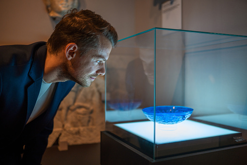 Mid adult man looking closely at the exhibited piece of glassware in a  case. Close up shot, side view of his face lit up by the display box lights.