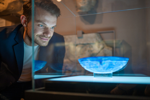 Mid adult attractive man having a close look at the exhibited glassware in a display box. Close up image, looking away, light from the spotlight reflecting on his face. Partly shot through glass.