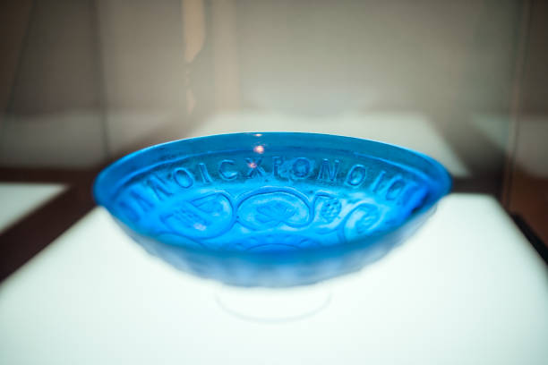 A close up of a beautiful shallow bowl made of blue transparent glass,  decorated with relief Greek letters. Found in the city of Ljubljana, exhibited as a part of Roman history collection. Gentle light coming through glass.