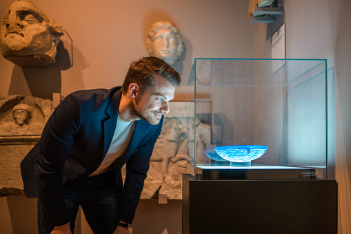 A man taking a close look at the glassware exhibited in a display glass box.  Waist up image, side view of  his face lit by bright reflection, looking away.