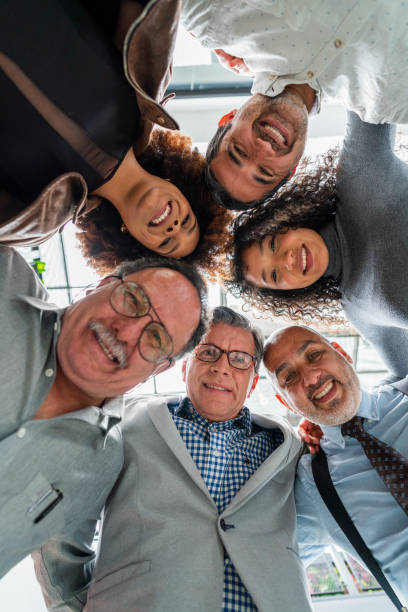 multiracial group of coworkers are photographed as they put their heads together and look down at the camera that portrays them multiracial group of formally dressed coworkers between the ages of 25 and 50 are inside the company they work for and are photographed as they put their heads together and look down at the camera that portrays them put down stock pictures, royalty-free photos & images