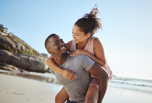 Love, piggy back and black couple on beach walking, smile and happy together, for bonding and outdoor. Romantic, man carry woman or loving on seaside vacation, holiday and romance for relationship.