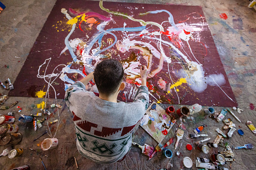 Rear view of the Caucasian young artist painting in his art studio. He is working with oil paints on a large canvas. Abstract art concepts.