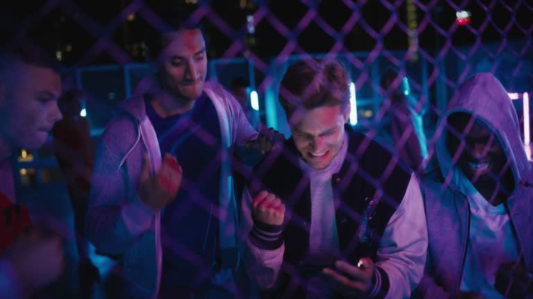 Group of Young Diverse Men Watching a Sports Game on Smartphone Device, Cheering for Their Favorite Team. Celebrating Goal, Betting or Lottery Win. Standing Outside in Urban Location with Neon Lights