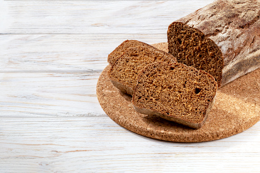 Loaf of dark bread with slices on a round board on a white wooden background