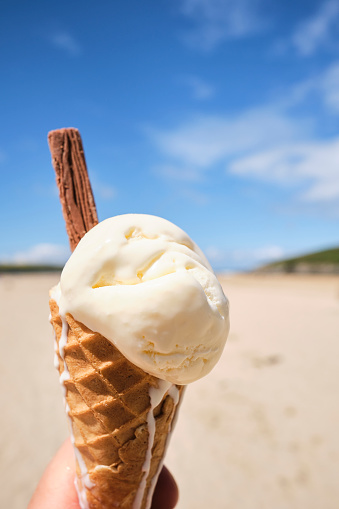 POV of a hand holding a melting vanilla Ice cream cone at Crantock beach, Newquay, Cornwall on a bright sunny day.