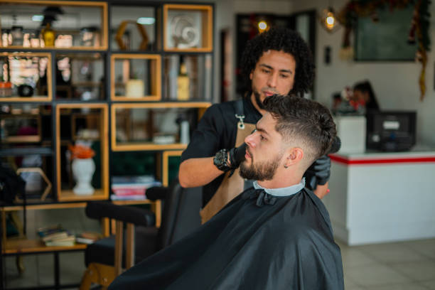 Professional stylist of a Latino man with afro curly hair, average age 30, dressed in his uniform, is in the barber shop he works for giving a haircut to a 25-year-old client