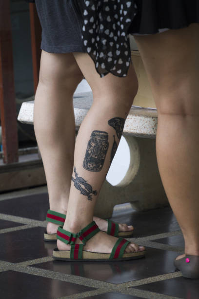 Male legs in with tattoos  of young man wearing shorts and sandals standing in group of young of young Thai people in Bangkok
