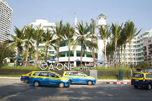 Hotels in Pattaya North and driving taxis in street. Local taxis are blue and yellow colored. In front of hotel in center are palm trees. Name of hotel is A-One