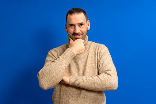 Photo of attractive bearded hispanic man wearing turtleneck thinking looking at copyspace hold hand chin isolated over blue color background