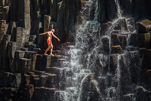 Caucasian man in swimsuit balancing near the falling water streams flowing on black volcanic stone cascades. Rochester Falls waterfall - popular tourist spot in Savanne district in Mauritius.