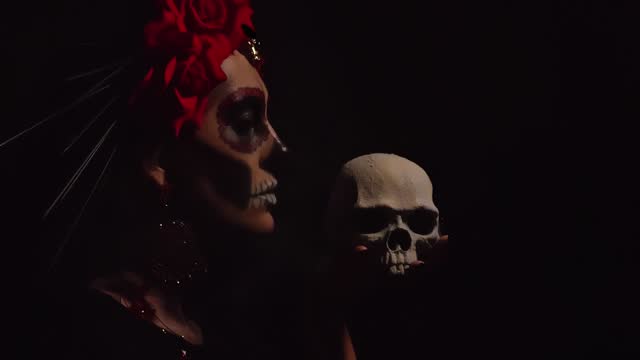 A woman with a pattern on her face in the style of the Goddess of Death with a sugar skull in front of her face on a black background blows white smoke on the scoop. Festival Day of the Dead