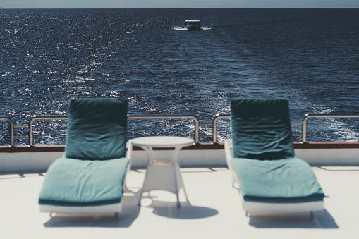 A wide-angle shot of a boat in selective focus on the background while in the foreground two recliners with blue cushions and a small coffee table, all on the upper deck of a luxury sailing yacht