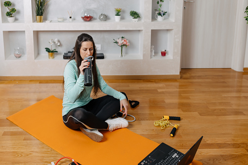 A mid-adult Caucasian woman is sitting on a yoga mat and drinking from a protein shaker.